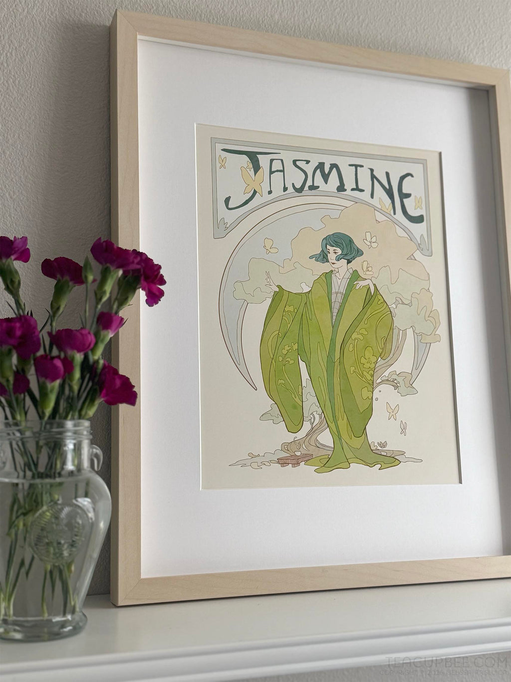 Framed art print of a woman in a green dress titled 'Jasmine' on a shelf with flowers.