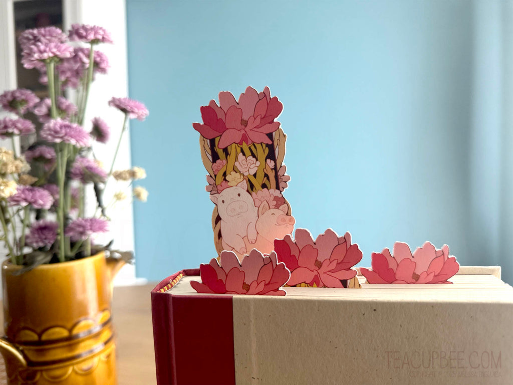 floral and piggy bookmark design sitting in a book next to a vase of flowers.