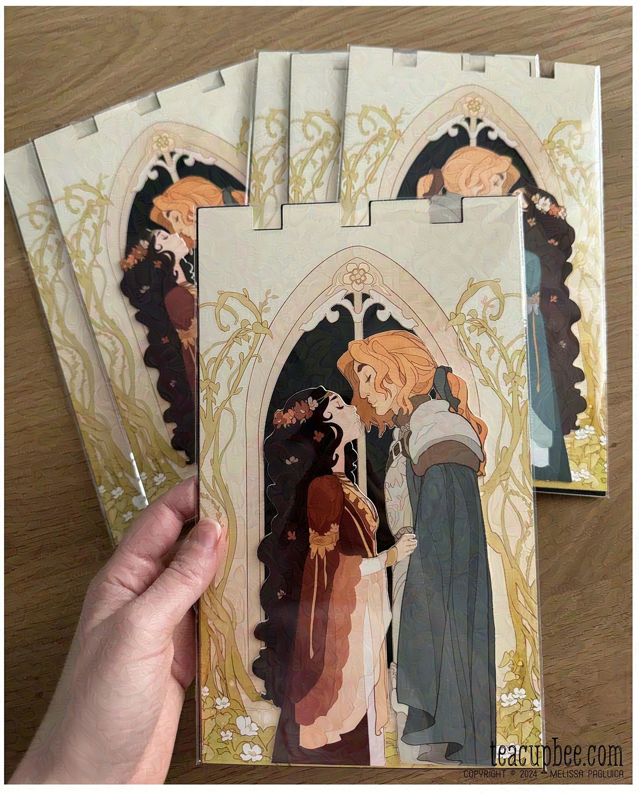 Set of illustrated bookmarks featuring two characters in a decorative frame, held by a hand on a wooden surface.